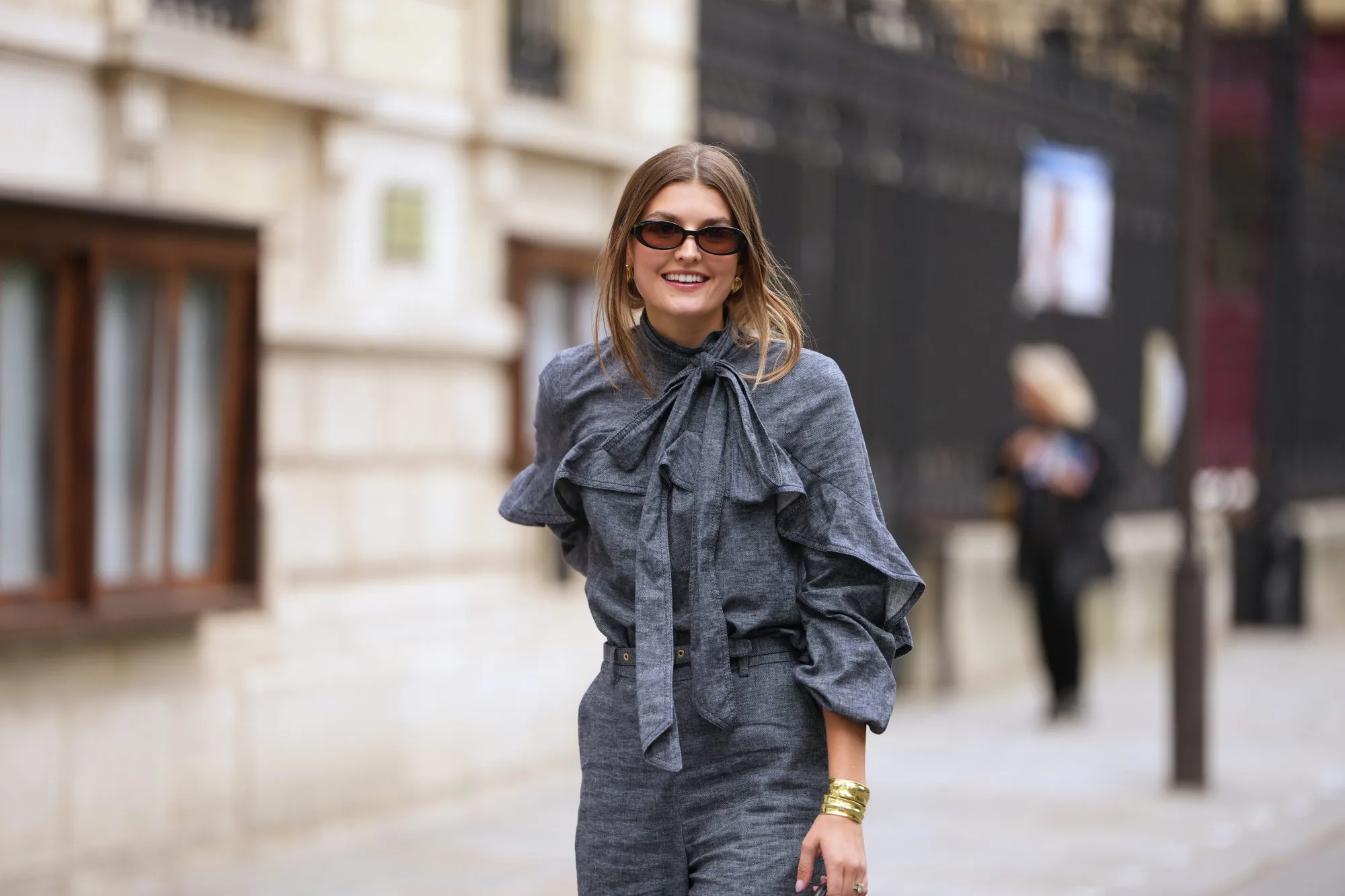 PARIS, FRANCE - OCTOBER 06: A guest wears black square sunglasses, a gray blue denim ruffled high neck / knot bow tie / long sleeves blouse, matching gray blue flared suit pants, outside Zimmermann, during Paris Fashion Week - Womenswear Spring Summer 2026, on October 06, 2025 in Paris, France (Photo by Edward Berthelot/Getty Images)