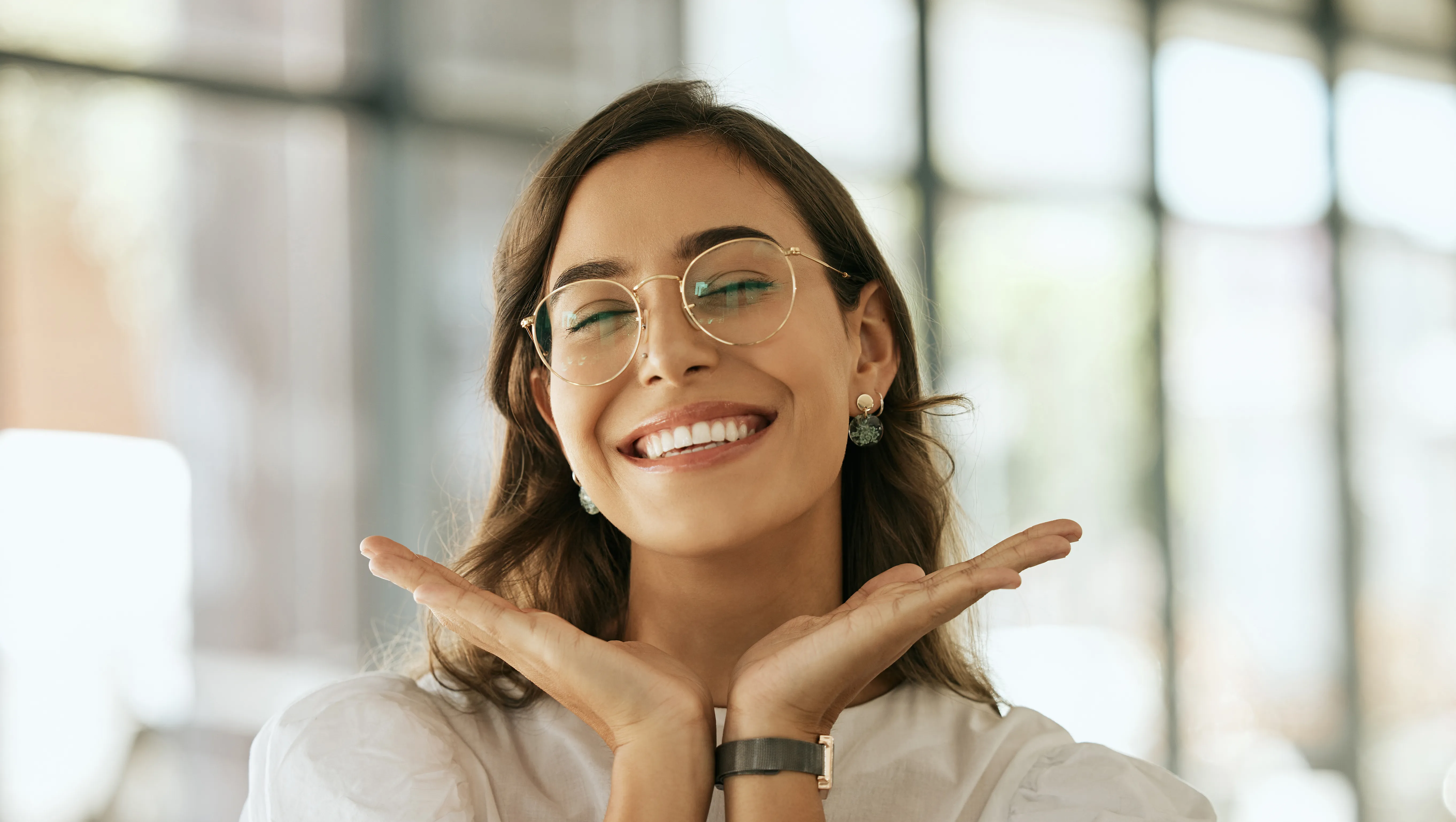 Cheerful business woman with glasses posing with her hands under her face showing her smile in an office. Playful hispanic female entrepreneur looking happy and excited at workplace