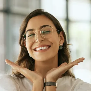 Cheerful business woman with glasses posing with her hands under her face showing her smile in an office. Playful hispanic female entrepreneur looking happy and excited at workplace