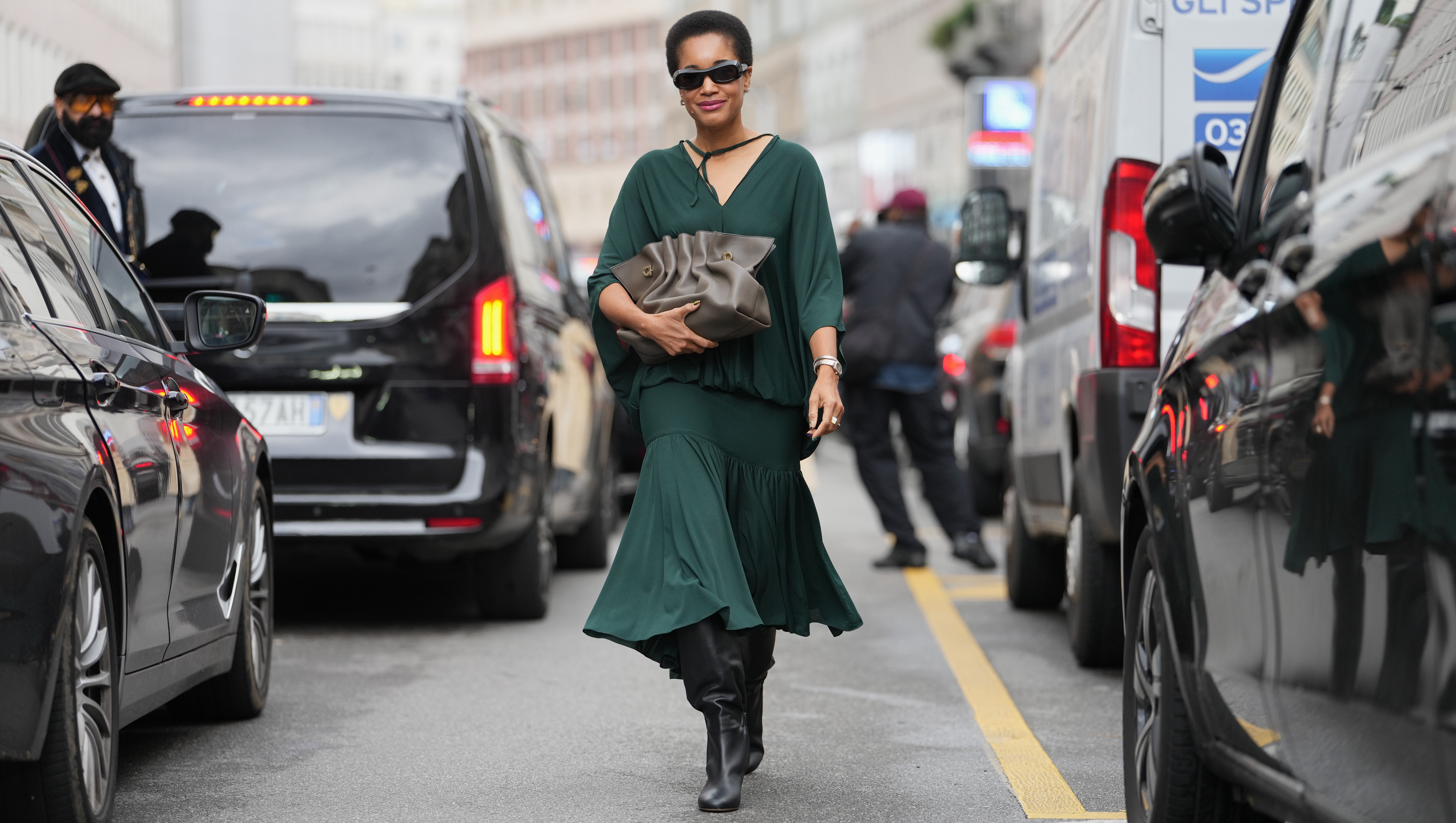 MILAN, ITALY - SEPTEMBER 27: Tamu McPherson wears a short natural black afro hairstyle, black rectangular sunglasses, small gold hoop earrings and a silver metal watch and bracelet, a taupe ruched leather pouch bag with gold-tone hardware handheld, a dark green flowing V-neck blouse with a tie at the neckline and dolman sleeves, a matching dark green gathered midi skirt with a tiered hem, black leather knee-high boots shoes with a block heel, outside Ferragamo, during Milan Fashion Week - Womenswear Spring/Summer 2026, on September 27, 2025 in Milan, Italy (Photo by Edward Berthelot/Getty Images)