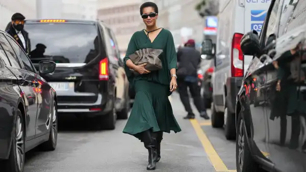 MILAN, ITALY - SEPTEMBER 27: Tamu McPherson wears a short natural black afro hairstyle, black rectangular sunglasses, small gold hoop earrings and a silver metal watch and bracelet, a taupe ruched leather pouch bag with gold-tone hardware handheld, a dark green flowing V-neck blouse with a tie at the neckline and dolman sleeves, a matching dark green gathered midi skirt with a tiered hem, black leather knee-high boots shoes with a block heel, outside Ferragamo, during Milan Fashion Week - Womenswear Spring/Summer 2026, on September 27, 2025 in Milan, Italy (Photo by Edward Berthelot/Getty Images)