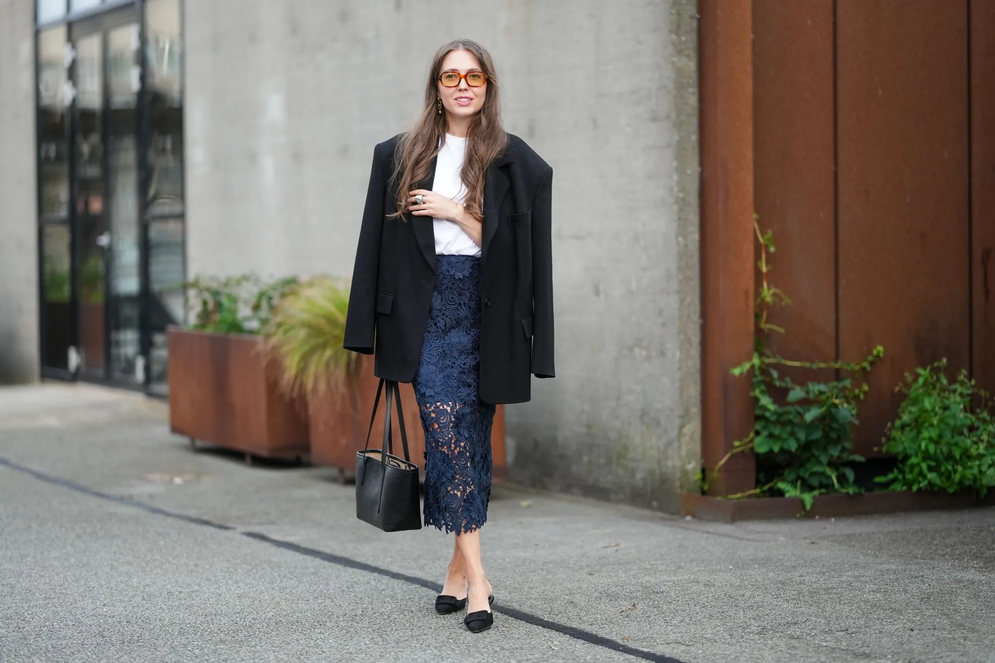 COPENHAGEN, DENMARK - AUGUST 07: A guest wears long wavy brown hair with a center part, oversized rectangular sunglasses with amber-tinted lenses, small gold drop earrings, an oversized black single-breasted blazer jacket with notched lapels and flap pockets draped over the shoulders, a white crew-neck T-shirt tucked into a navy floral lace pencil midi skirt with partial sheerness through the legs and a scalloped hem, a black structured leather tote with twin top handles, stacked rings including a chunky silver-toned ring, pale nude nail polish, and black slingback pumps with low block heels and a bow at the toe, walking, outside Rotate, during Copenhagen Fashion Week, on August 07, 2025 in Copenhagen, Denmark (Photo by Edward Berthelot/Getty Images)