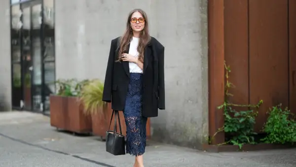 COPENHAGEN, DENMARK - AUGUST 07: A guest wears long wavy brown hair with a center part, oversized rectangular sunglasses with amber-tinted lenses, small gold drop earrings, an oversized black single-breasted blazer jacket with notched lapels and flap pockets draped over the shoulders, a white crew-neck T-shirt tucked into a navy floral lace pencil midi skirt with partial sheerness through the legs and a scalloped hem, a black structured leather tote with twin top handles, stacked rings including a chunky silver-toned ring, pale nude nail polish, and black slingback pumps with low block heels and a bow at the toe, walking, outside Rotate, during Copenhagen Fashion Week, on August 07, 2025 in Copenhagen, Denmark (Photo by Edward Berthelot/Getty Images)
