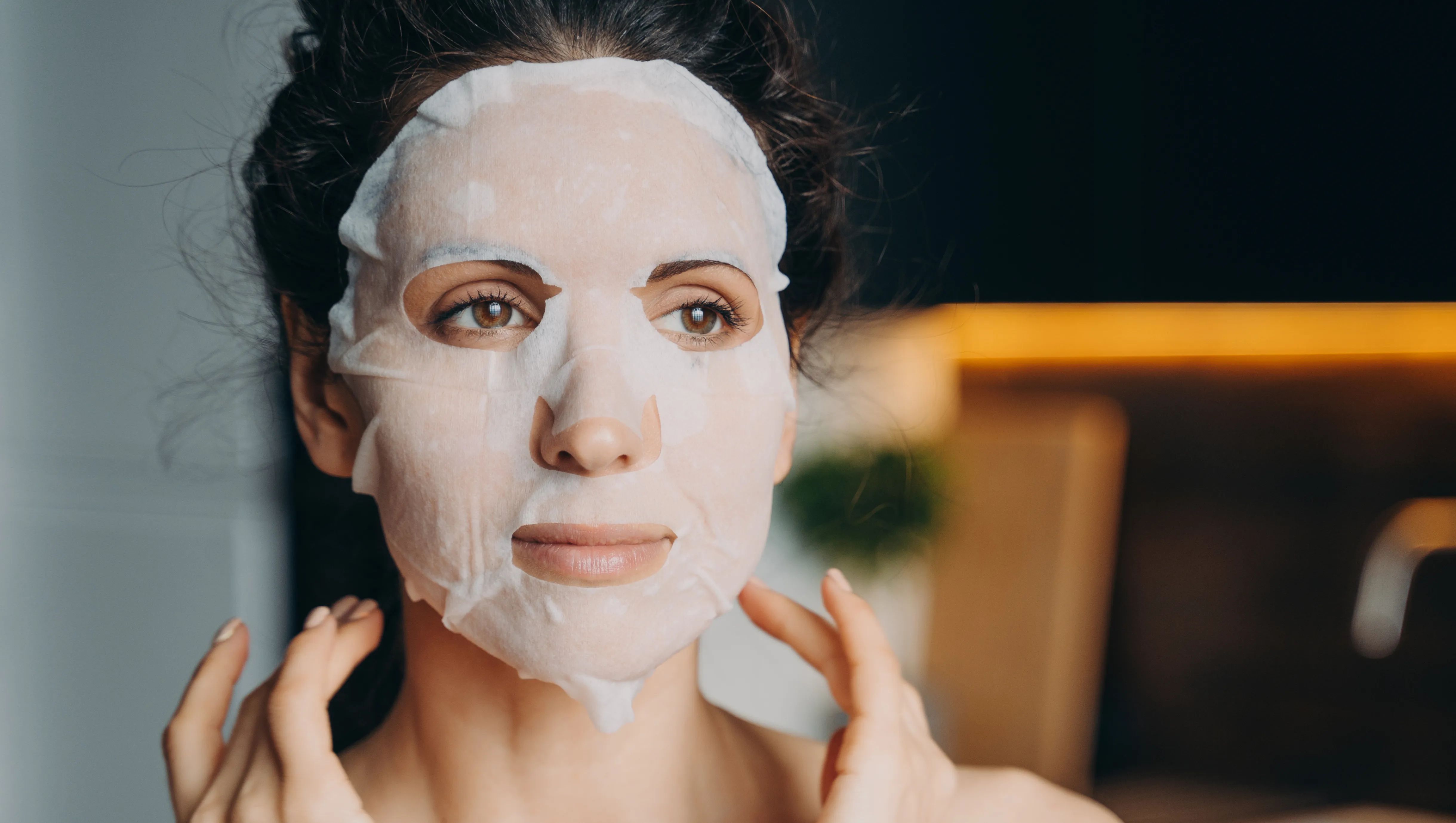 Young woman with bare shoulders applies moisturizing rejuvenation collagen facial sheet mask to improve complexion, for face skin treatment at home. Skincare, daily beauty routine, self-care.