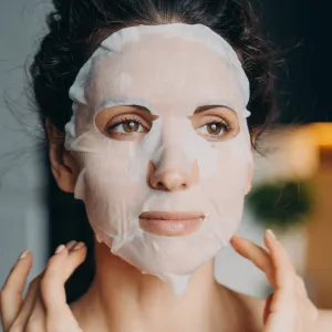 Young woman with bare shoulders applies moisturizing rejuvenation collagen facial sheet mask to improve complexion, for face skin treatment at home. Skincare, daily beauty routine, self-care.