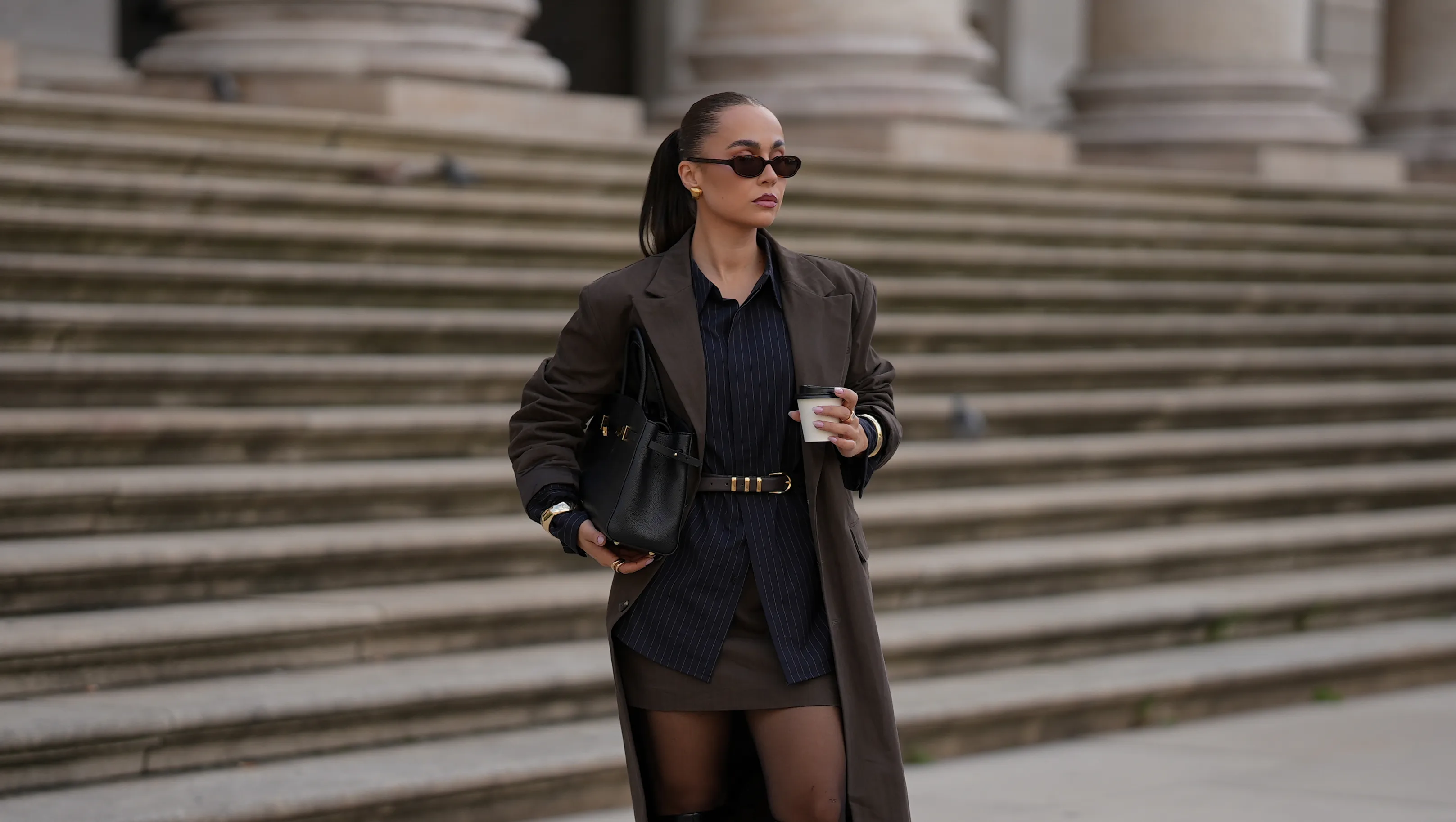 MUNICH, GERMANY - SEPTEMBER 16: Anna Wein is seen wearing a long brown coat with long sleeves and pinstripe details from Lounge; a dark pinstriped shirt with black buttons; a short brown skirt with subtle pinstripes from Lounge; sheer black tights; tall black leather heeled boots with a chunky heel and a square toe from H&M; a structured black leather handbag with gold hardware; a black leather belt, tied around the waist, with a golden fastener; slim dark sunglasses with brown tinted lenses from Corlin Eyewear; subtle golden metallic jewelry featuring rings and bracelets from Mango; her brown hair is styled in a sleek ponytail on September 16, 2025 in Munich, Germany. (Photo by Moritz Scholz/Getty Images)