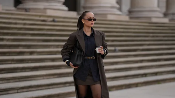 MUNICH, GERMANY - SEPTEMBER 16: Anna Wein is seen wearing a long brown coat with long sleeves and pinstripe details from Lounge; a dark pinstriped shirt with black buttons; a short brown skirt with subtle pinstripes from Lounge; sheer black tights; tall black leather heeled boots with a chunky heel and a square toe from H&M; a structured black leather handbag with gold hardware; a black leather belt, tied around the waist, with a golden fastener; slim dark sunglasses with brown tinted lenses from Corlin Eyewear; subtle golden metallic jewelry featuring rings and bracelets from Mango; her brown hair is styled in a sleek ponytail on September 16, 2025 in Munich, Germany. (Photo by Moritz Scholz/Getty Images)