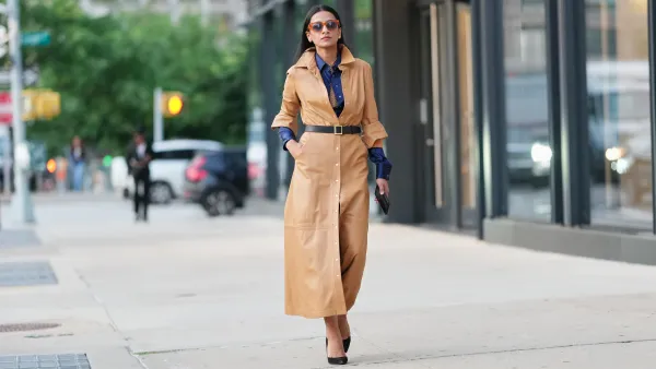 NEW YORK, NEW YORK - SEPTEMBER 12: A guest wears long straight dark hair parted in the center, tortoiseshell rectangular sunglasses with brown lenses, a tan leather long belted button-front shirt dress with a shirt collar and visible silver snap buttons and a center-front slit, a black leather belt with a square gold buckle cinching the waist, a navy blue button-up shirt with visible navy cuffs and a brown knit tie, and black pointed-toe pumps with stiletto heels, outside Sergio Hudson, during New York Fashion Week, on September 12, 2025 in New York, New York (Photo by Edward Berthelot/Getty Images)