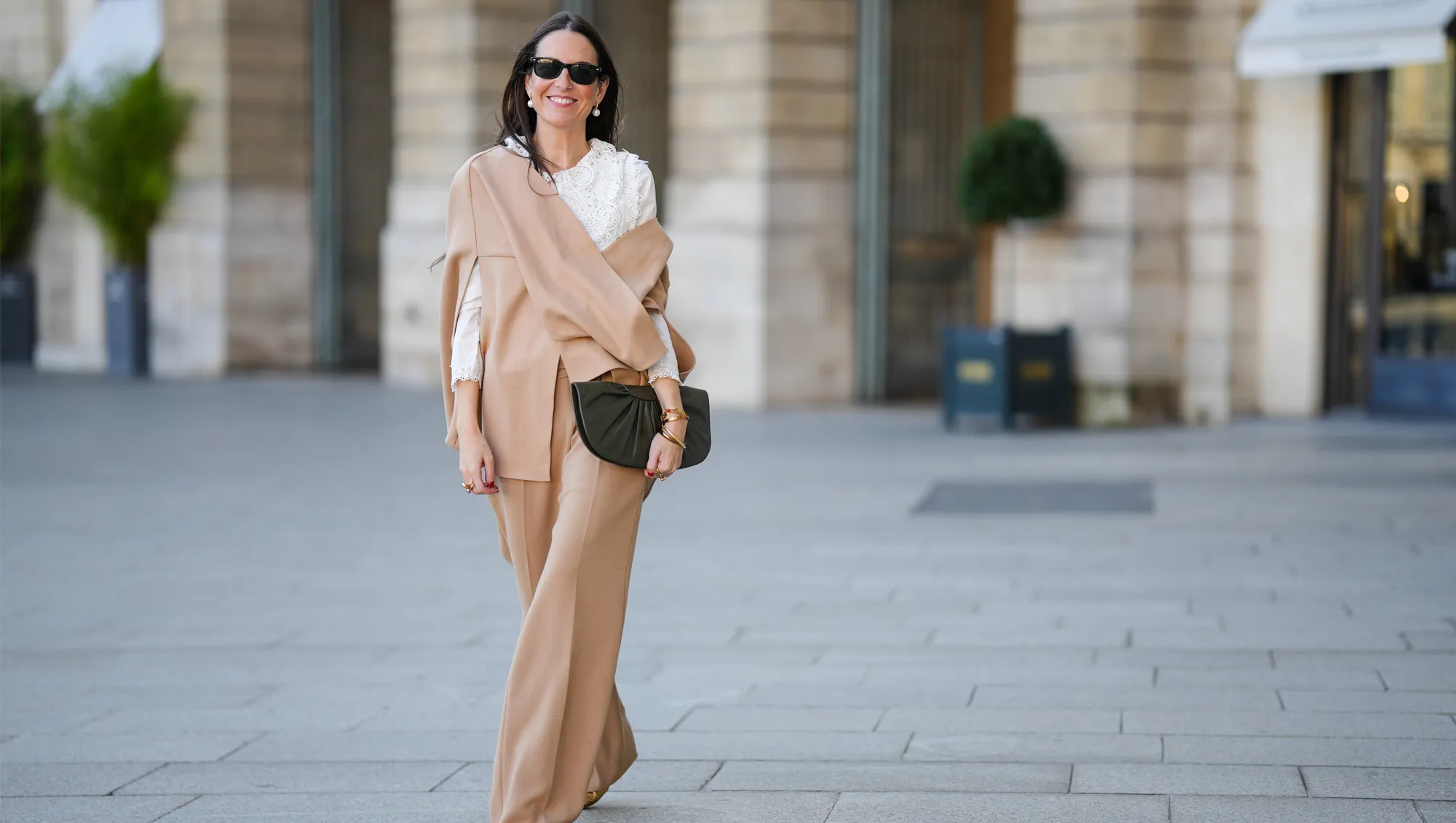 Alba Garavito Torre wears black sunglasses from Ray Ban, white large pearls earrings, a white shirt with cut-out lace pattern large collar, a beige felt / wool oversized cloak, matching beige wool / felt palazzo pants, a black shiny leather ruffled handbag, a gold Love bracelet from Cartier, gold bracelet, gold rings, brown leather pointed pumps heels shoes, during a street style fashion photo session, on October 28, 2021 in Paris, France.