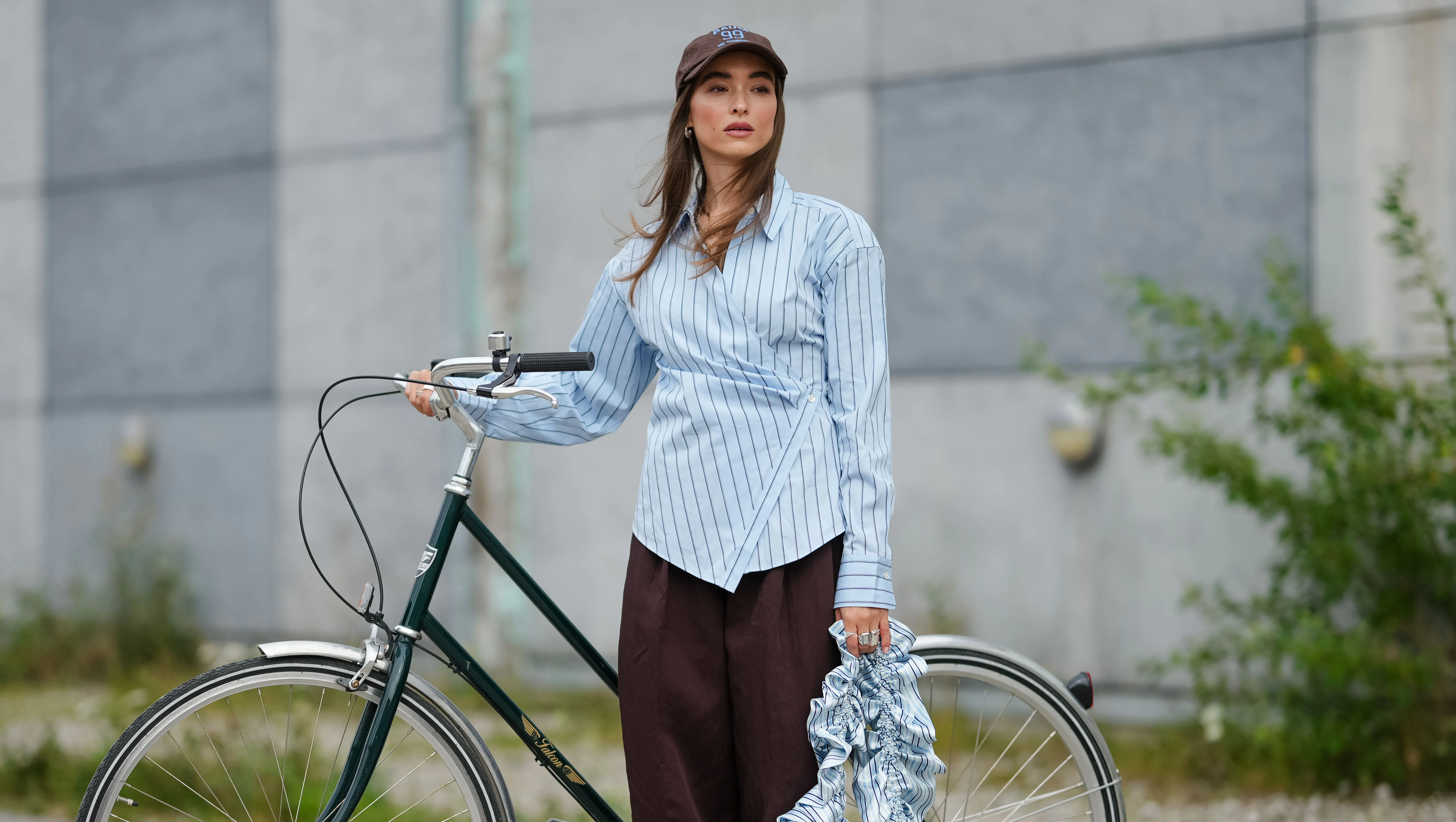 COPENHAGEN, DENMARK - AUGUST 07: A guest wears a brown baseball cap with embroidered details. Hair is styled straight and loose. The outfit features a light blue button-up shirt with vertical dark blue stripes, designed with an asymmetrical wrap detail. Paired with wide-legged, dark brown trousers. Footwear consists of blue sandals. Accessories include a ruffled, striped bag in shades of blue and white, outside Marimekko, during Copenhagen Fashion Week, on August 07, 2025 in Copenhagen, Denmark (Photo by Edward Berthelot/Getty Images)