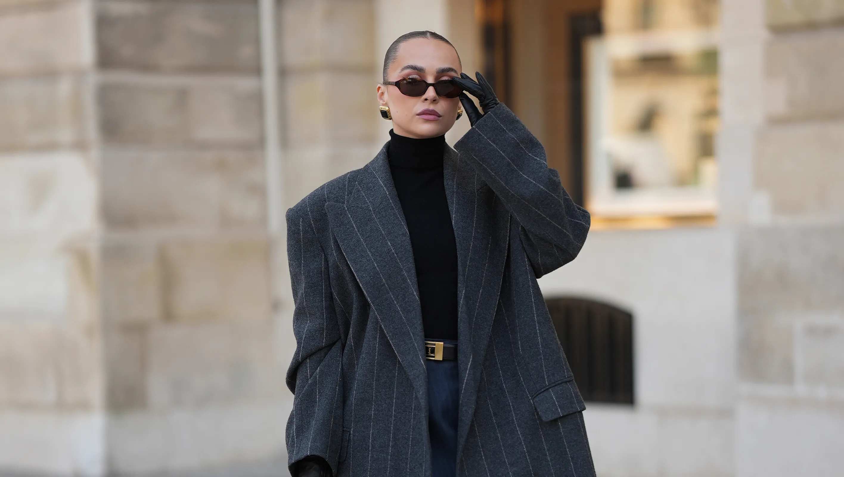 PARIS, FRANCE - OCTOBER 1: Anna Wein is wearing grey pinstripe long coat by Bazhane over a black turtleneck from H&M, a navy pinstripe mini skirt from H&M, a black belt by Yves Saint Laurent and knee-high black leather boots from H&M; she accessorizes with a black Chanel handbag with gold chain detail, black leather gloves, gold hoop earrings from H&M, and dark rectangular sunglasses from Corlin Eyewear; her hair is styled in a sleek low bun; the look is captured outside as part of Paris Fashion Week on October 1, 2025 in Paris, France. (Photo by Moritz Scholz/Getty Images)