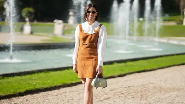 PARIS, FRANCE - OCTOBER 5: Sabrina Belnick is seen wearing a full Look from Celine including a brown suede pinafore-style mini dress with front pockets, seam-lines and gold buckle details; a white long-sleeve blouse with a bow-tie neckline and slightly puffed sleeves with cuffs; brown leather ankle boots with a sturdy heel and minimal branding; the Teen Lola, light clay crocodile-embossed handbag with gold clasp closure and a logo-patterned white-grey fabric wrapped around the top handle; slim round sunglasses with metallic frames; her brown hair is worn in a straight fringed bob, with a middle part during the Celine Womenswear Spring Summer 2026 Show as part of Paris Fashion Week on October 5, 2025 in Paris, France. (Photo by Moritz Scholz/Getty Images)