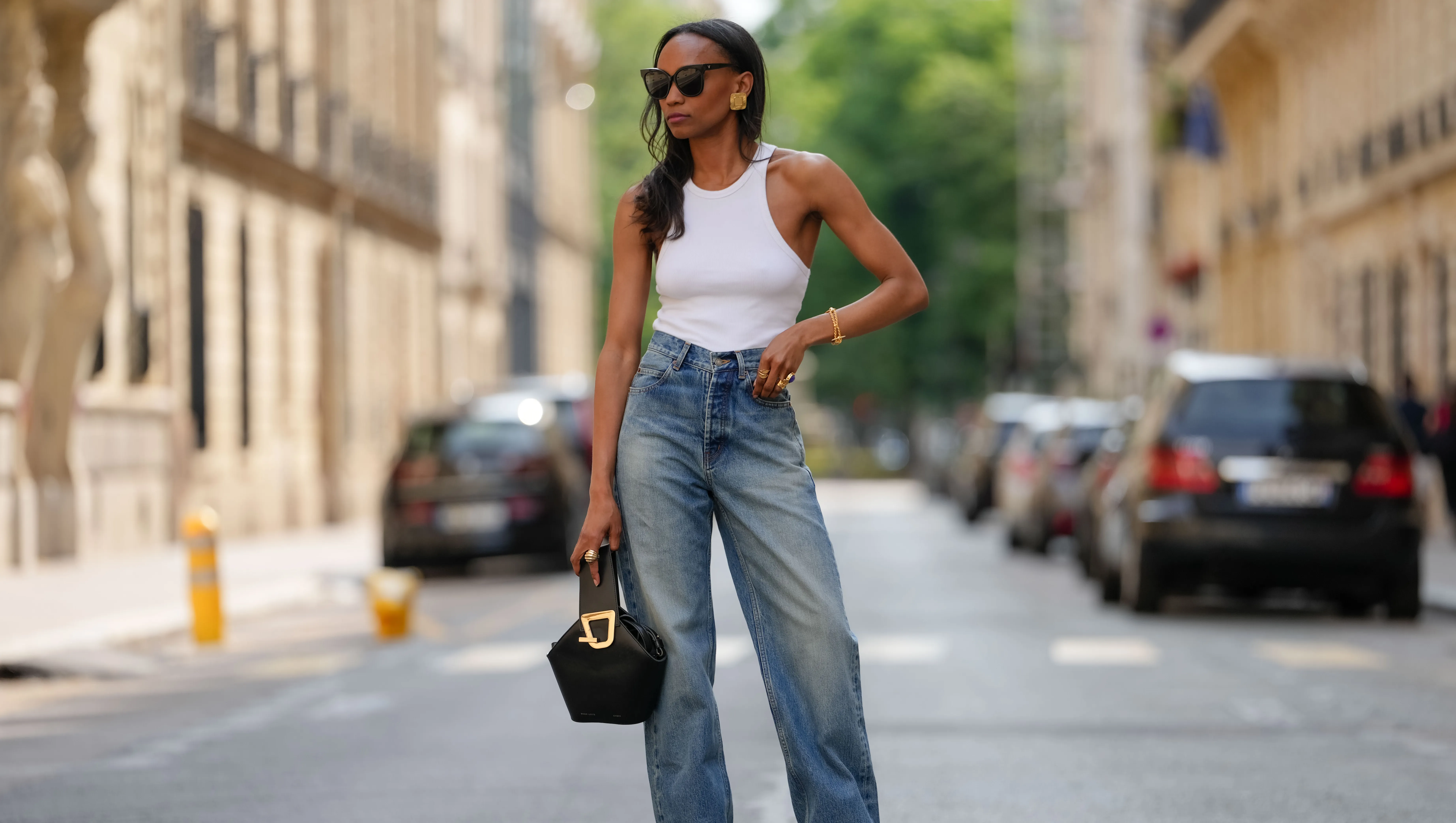 PARIS, FRANCE - JUNE 14: Emilie Joseph wears a white ribbed tank top from Anine Bing paired with high-waisted blue denim jeans from YSL. The jeans feature a relaxed, wide-leg cut. Accessories include large gold hoop earrings, a gold bracelet, and black cat-eye sunglasses. A black structured handbag with a gold buckle detail is carried. Footwear consists of black pointed-toe shoes from YSL Saint Laurent with a visible gold accent, during a street style fashion photo session, on June 14, 2025 in Paris, France (Photo by Edward Berthelot/Getty Images)