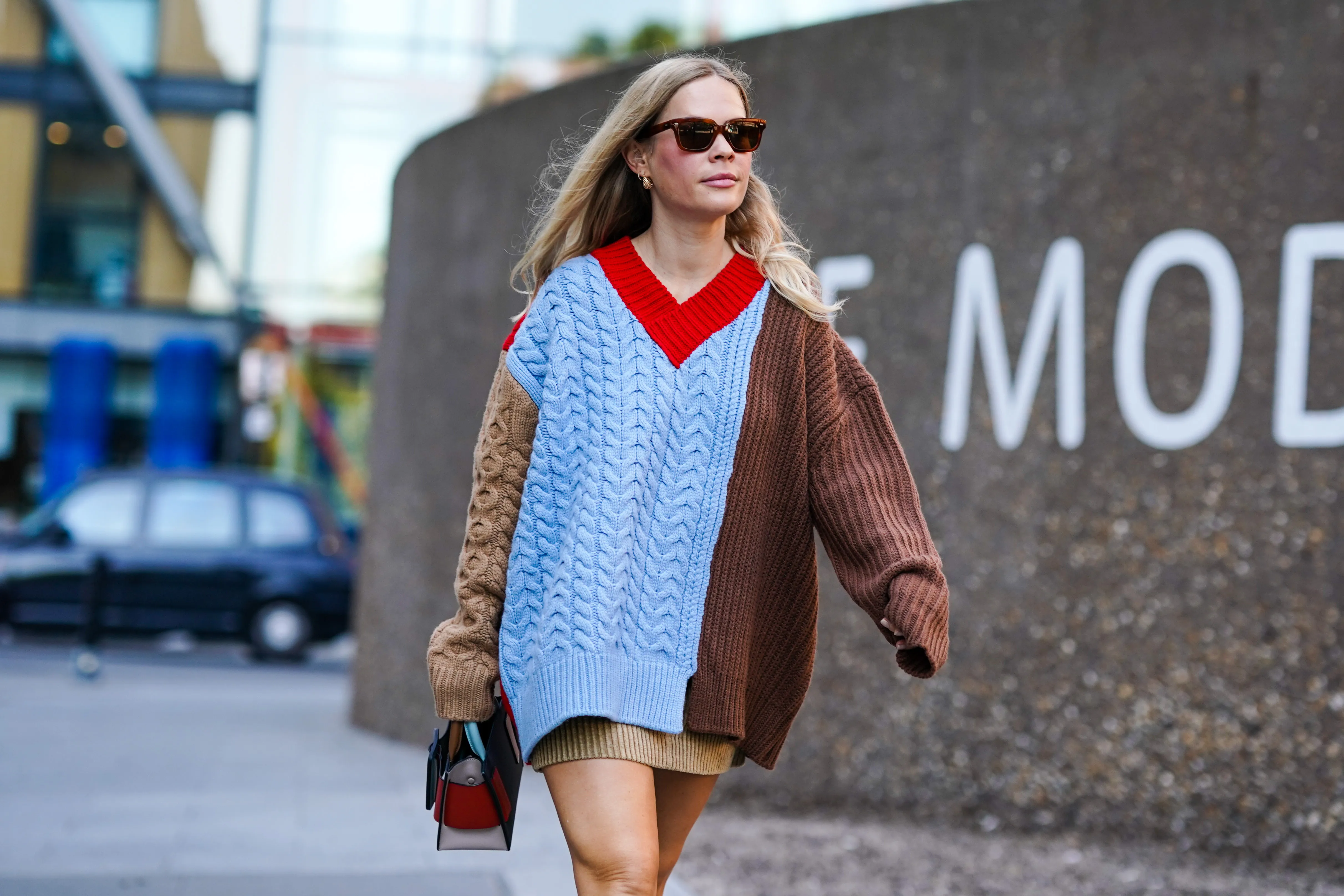 A guest wears sunglasses, earrings, a brown and blue knitted wool pullover/dress with red v-neck, a bag, during London Fashion Week September 2019 on September 14, 2019 in London, England.