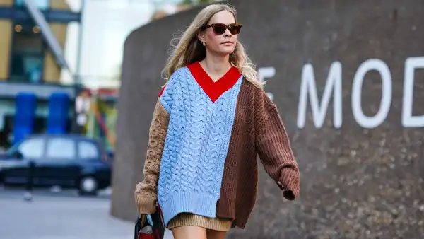 A guest wears sunglasses, earrings, a brown and blue knitted wool pullover/dress with red v-neck, a bag, during London Fashion Week September 2019 on September 14, 2019 in London, England.