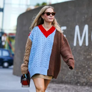 A guest wears sunglasses, earrings, a brown and blue knitted wool pullover/dress with red v-neck, a bag, during London Fashion Week September 2019 on September 14, 2019 in London, England.