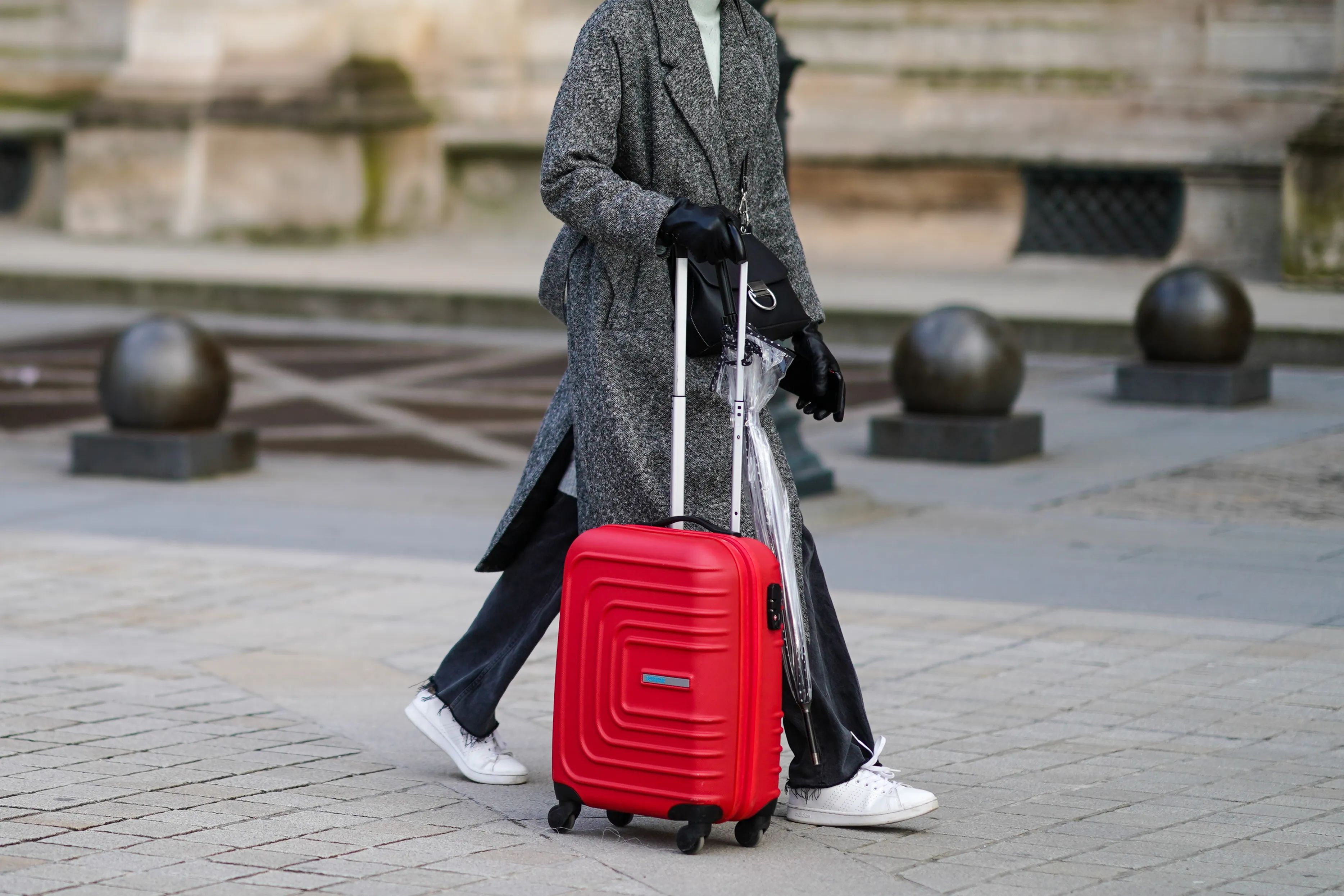 PARIS, FRANCE - JANUARY 17: A passerby wears a white woven turtleneck wool pullover, a gray oversized long coat, black denim jeans ripped pants, white leather laces sneakers, black shiny leather gloves, a black shiny leather crossbody bag, a transparent umbrella, a red large suitcase, on January 17, 2021 in Paris, France. (Photo by Edward Berthelot/Getty Images)
