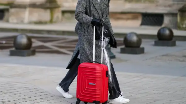 PARIS, FRANCE - JANUARY 17: A passerby wears a white woven turtleneck wool pullover, a gray oversized long coat, black denim jeans ripped pants, white leather laces sneakers, black shiny leather gloves, a black shiny leather crossbody bag, a transparent umbrella, a red large suitcase, on January 17, 2021 in Paris, France. (Photo by Edward Berthelot/Getty Images)