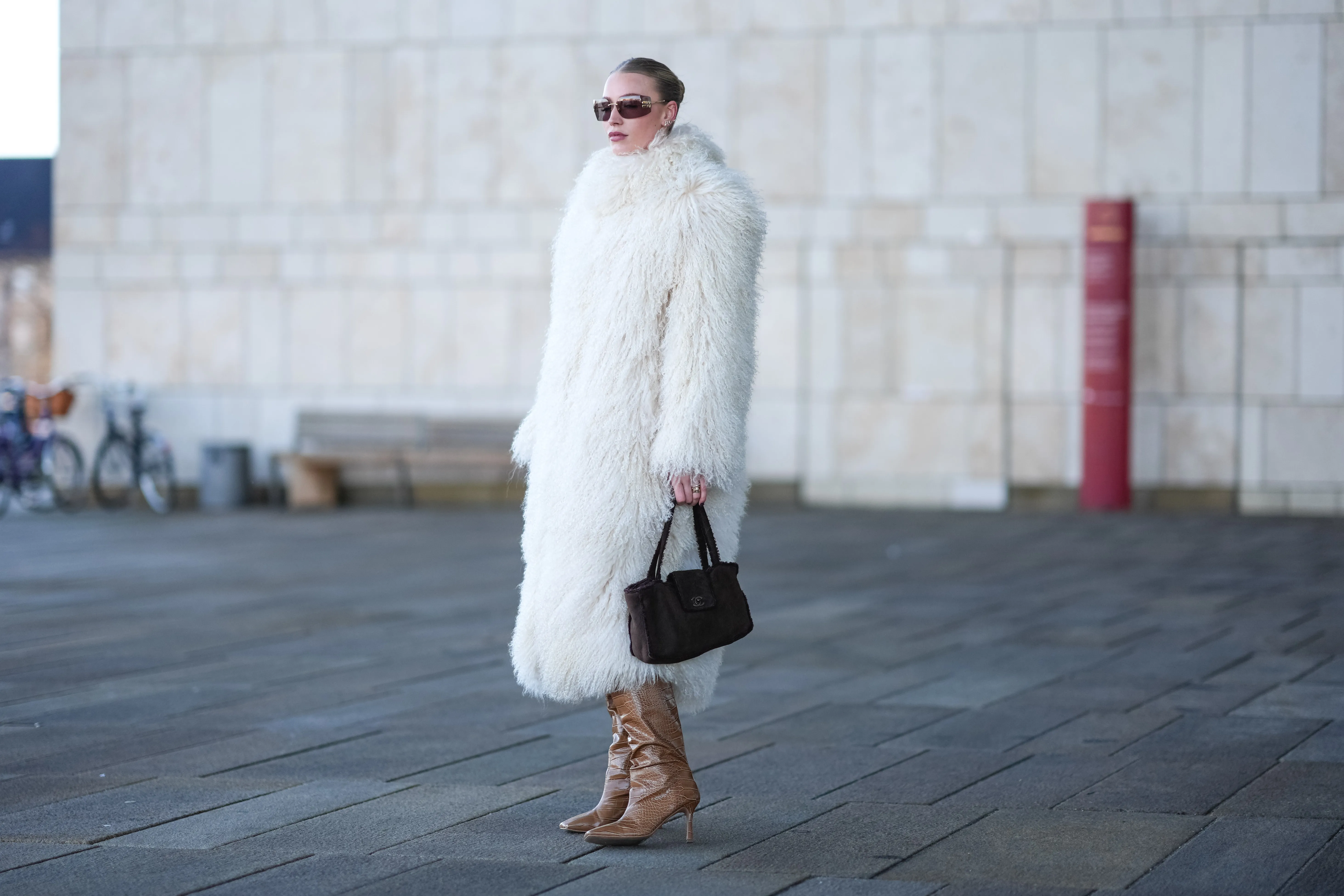 A guest wears Miu Miu sunglasses, a white oversized fluffy long faux fur coat , a Chanel bag, high heels pointed boots in brown leather, dark pink lipstick, outside OperaSport, during the Copenhagen Fashion Week AW24 on January 31, 2024 in Copenhagen, Denmark.