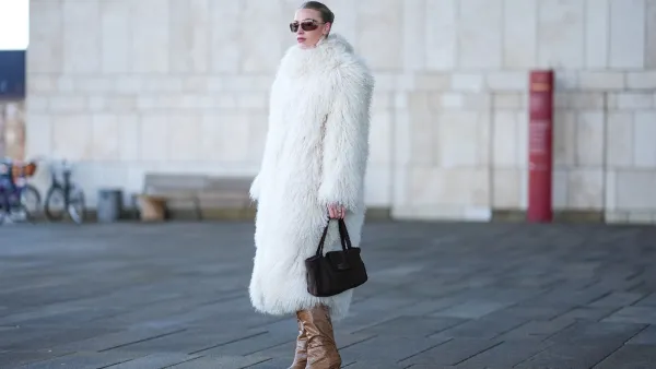 A guest wears Miu Miu sunglasses, a white oversized fluffy long faux fur coat , a Chanel bag, high heels pointed boots in brown leather, dark pink lipstick, outside OperaSport, during the Copenhagen Fashion Week AW24 on January 31, 2024 in Copenhagen, Denmark.