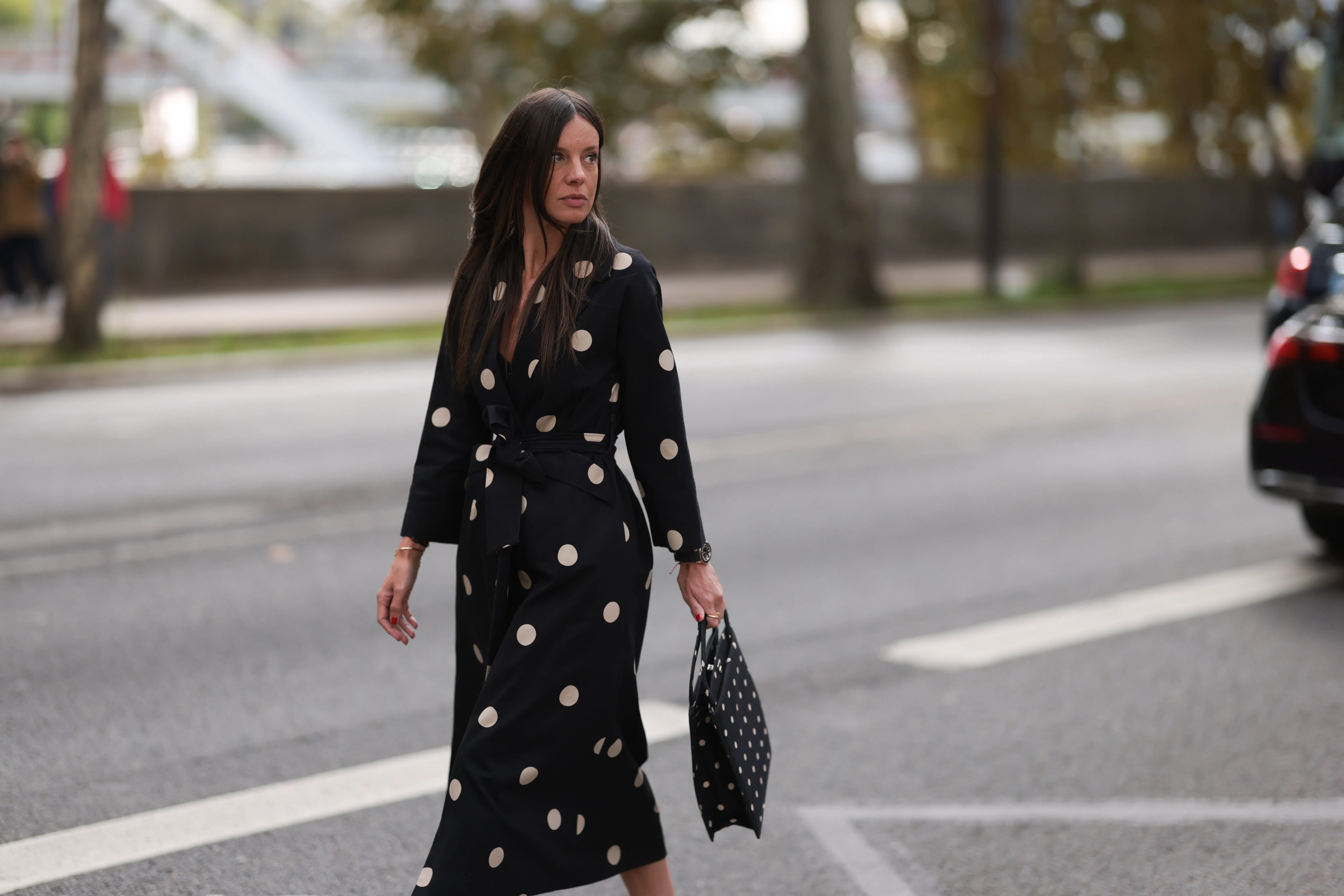 A Fashion Week guest is seen wearing round diamond stud earrings; a long-sleeved black midi dress with white polka dot pattern, notched lapels, and a tie belt; a gold bangle; a rose gold and black watch from Rolex; a gold ring; and a rectangular black handbag with a white polka dot pattern before the Elie Saab Womenswear Spring/Summer 2025 Show as part of Paris Fashion Week on September 28, 2024 in Paris, France.