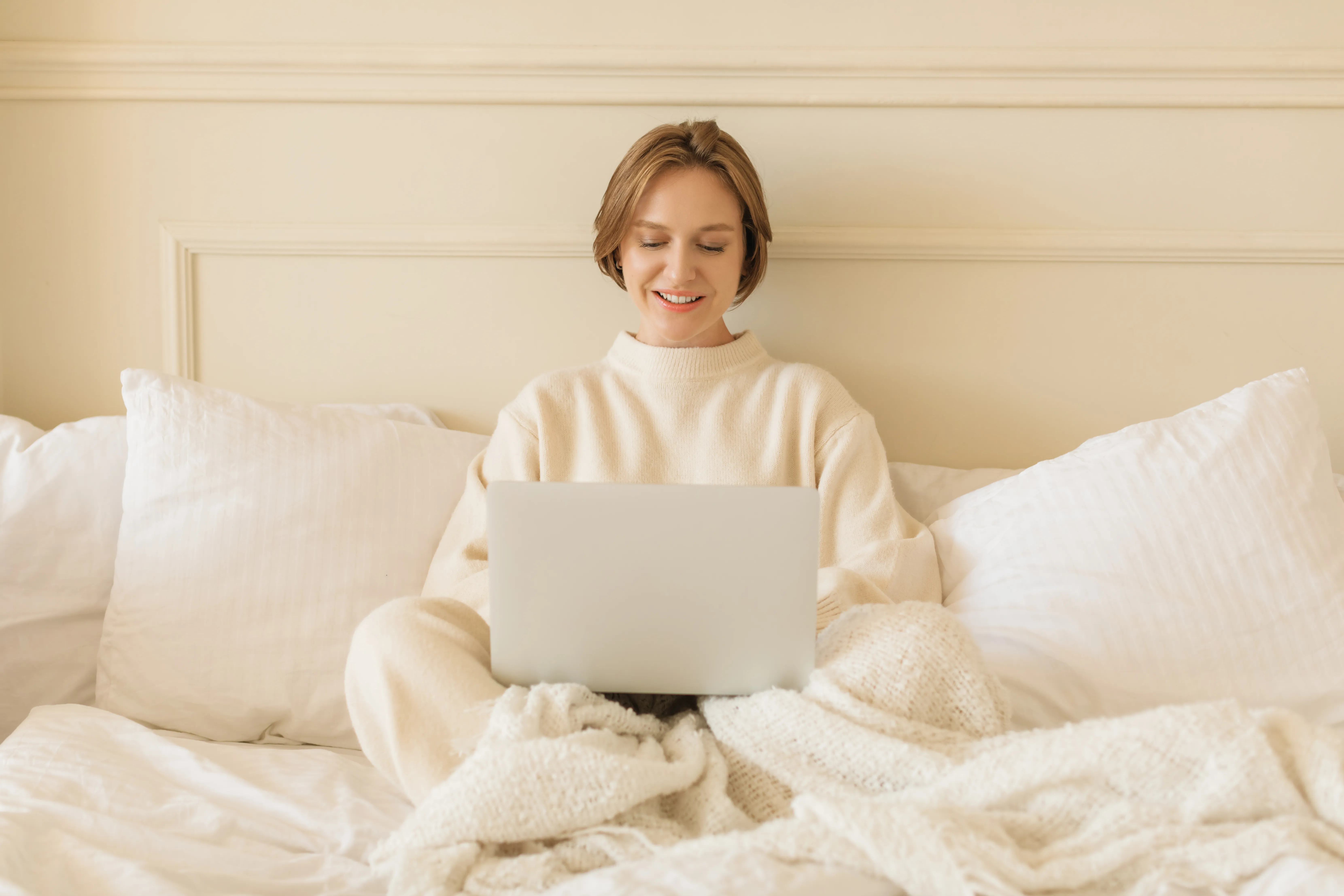 Smiling young woman in casual loungewear sitting on her bed working on her laptop computer
