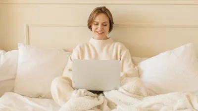 Smiling young woman in casual loungewear sitting on her bed working on her laptop computer
