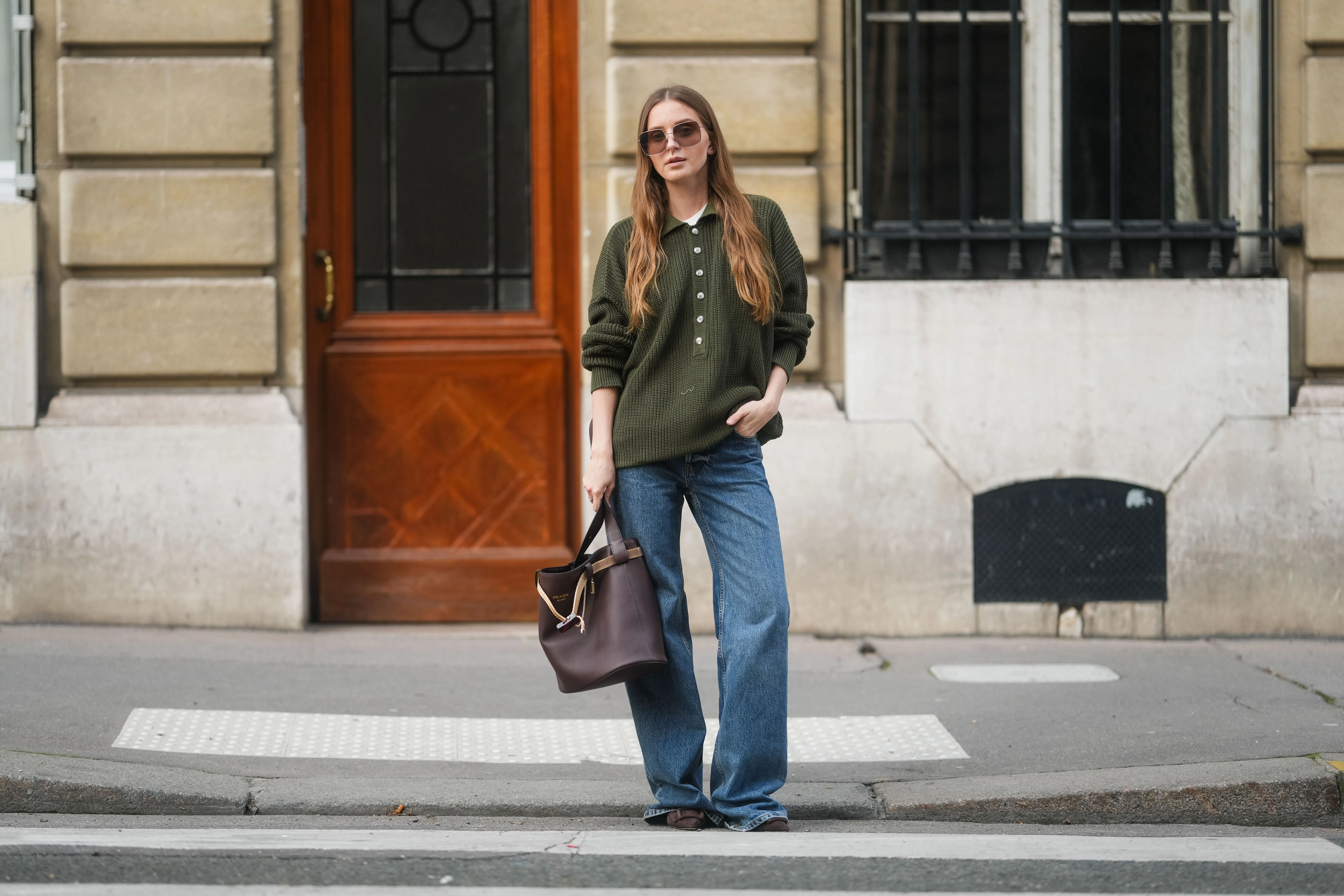 PARIS, FRANCE - MARCH 25: Diane Batoukina is seen wearing long straight brown hair, square Dior sunglasses, an oversized khaki green knitted polo sweater with mother-of-pearl buttons by By Aylin Koenig, blue wide-leg Anine Bing jeans, and carrying a brown Prada bucket bag with a gold chain handle, during a street style fashion photo session on March 25, 2025 in Paris, France. (Photo by Edward Berthelot/Getty Images)