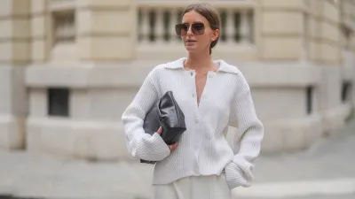 Diane Batoukina wears a white ribbed knit cardigan with a wide collar and oversized sleeves by Lovechild 1979, paired with high-waisted ivory trousers by Herskind. A dark brown leather clutch bag by St. Agni is held under one arm. Oversized square Dior sunglasses with brown gradient lenses are worn. Hair is parted at the center and styled in a sleek low bun. Natural makeup with even skin and soft pink lips is visible. Mid shot, during a street style fashion photo session, on April 17, 2025 in Paris, France.