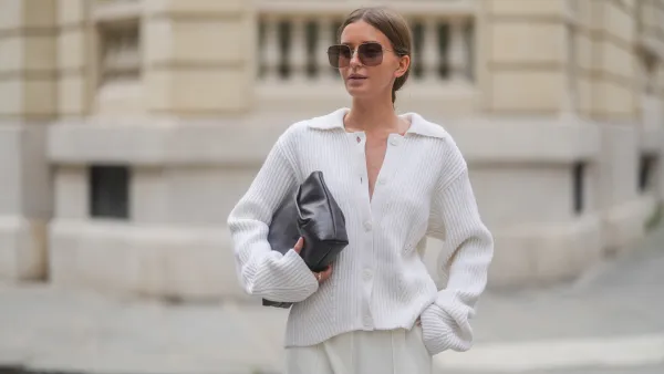 Diane Batoukina wears a white ribbed knit cardigan with a wide collar and oversized sleeves by Lovechild 1979, paired with high-waisted ivory trousers by Herskind. A dark brown leather clutch bag by St. Agni is held under one arm. Oversized square Dior sunglasses with brown gradient lenses are worn. Hair is parted at the center and styled in a sleek low bun. Natural makeup with even skin and soft pink lips is visible. Mid shot, during a street style fashion photo session, on April 17, 2025 in Paris, France.