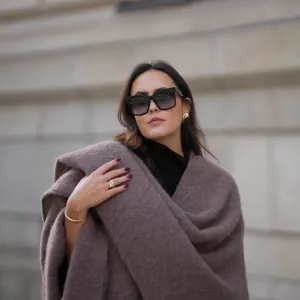 Laura Noltemeyer is seen wearing an oversized fringed wool poncho in a mauve tone with a textured, fluffy finish, fringes at the hem and a gold pin closure element called „Poncho COZY“ by Aylin Koenig; black fine-knit turtleneck sweater with slim-fitting silhouette by Aylin Koenig; among her jewelry is a white gold „LOVE“ bracelet by Cartier; oversized square black acetate sunglasses with ombré tinted lenses by Céline; large gold statement earrings with a ruffled surface and a green stone element by Mango; her dark brown hair is styled in loose waves with a center part on November 13, 2025 in Hamburg, Germany.