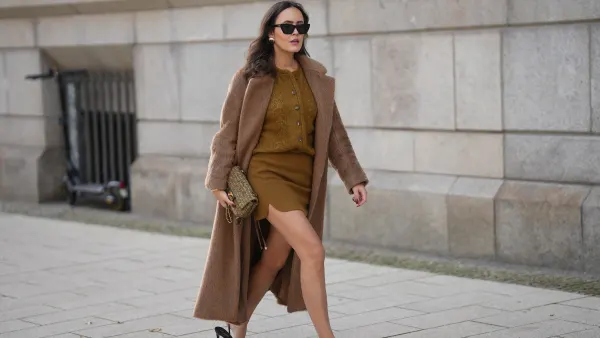 Laura Noltemeyer is seen wearing a a long camel-colored brushed wool coat with a wrap silhouette and integrated belt by Max Mara; a mustard-colored knit cardigan with a brushed cable-knit pattern and front button fastening by Sézane; a mustard yellow brownish mini skirt in a soft wool fabric by Sézane; black patent leather slingback pumps with a sharply pointed toe and silver hardware accents by Yves Saint Laurent; a small quilted tweed flap bag in a khaki Color with woven fabric material with a gold chain strap and CC clasp by Chanel; gold statement earrings with a ruffled surface and a green stone element by Mango; black cat eye sunglasses with an acetate frame by Yves Saint Laurent; her brown hair is styled in loose waves with a center part on November 13, 2025 in Hamburg, Germany.