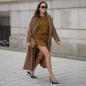 Laura Noltemeyer is seen wearing a a long camel-colored brushed wool coat with a wrap silhouette and integrated belt by Max Mara; a mustard-colored knit cardigan with a brushed cable-knit pattern and front button fastening by Sézane; a mustard yellow brownish mini skirt in a soft wool fabric by Sézane; black patent leather slingback pumps with a sharply pointed toe and silver hardware accents by Yves Saint Laurent; a small quilted tweed flap bag in a khaki Color with woven fabric material with a gold chain strap and CC clasp by Chanel; gold statement earrings with a ruffled surface and a green stone element by Mango; black cat eye sunglasses with an acetate frame by Yves Saint Laurent; her brown hair is styled in loose waves with a center part on November 13, 2025 in Hamburg, Germany.