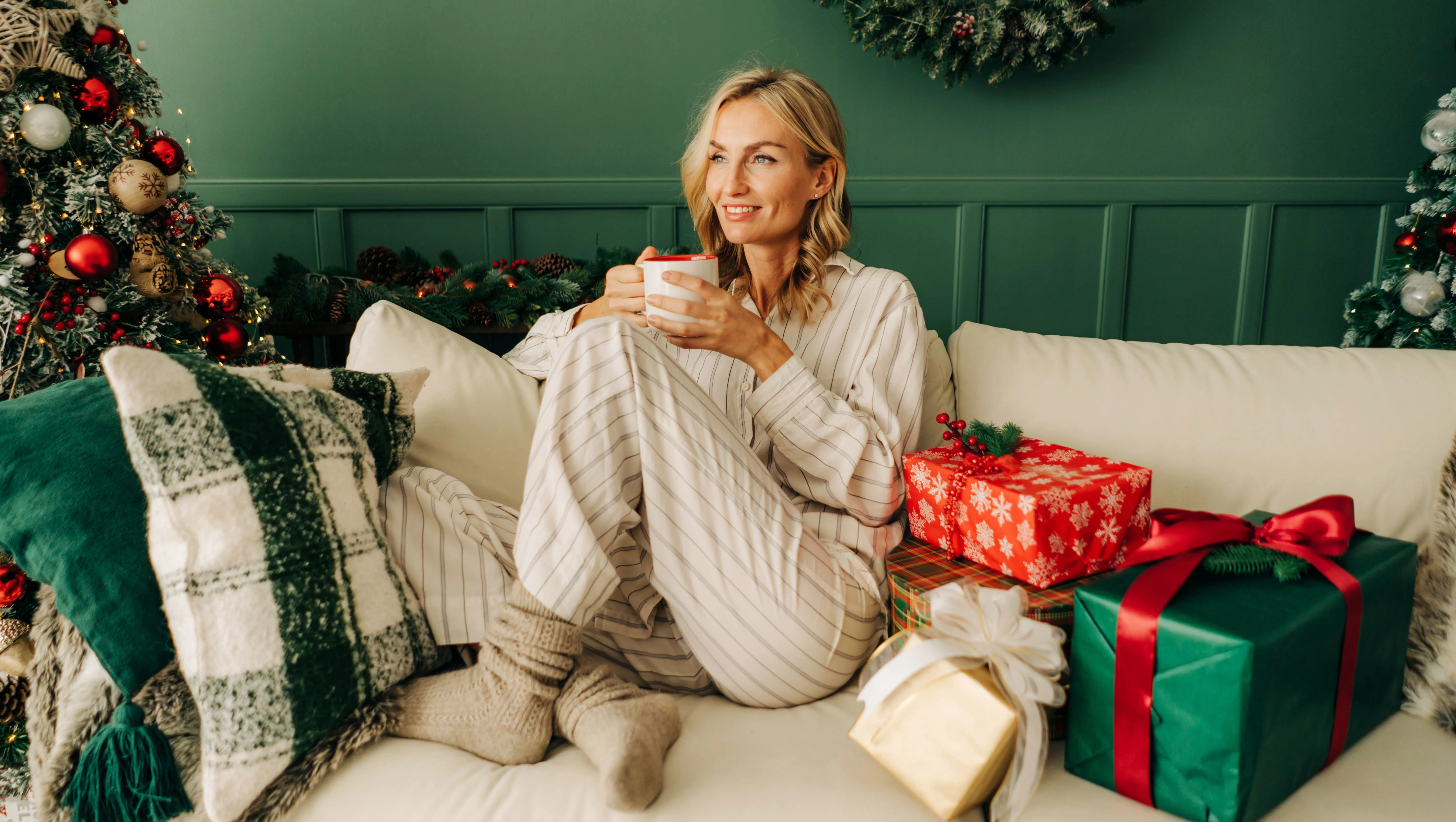 A beautiful blonde woman is sitting on the sofa in the Christmas living room and drinking coffee.