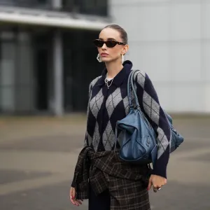 DUESSELDORF, GERMANY - OCTOBER 14: Evi Wave is seen wearing a navy blue and grey argyle knit H&M cardigan with a collared neckline, front button closure with tonal buttons and ribbed cuffs; some delicate chain and ball chain necklaces; a plaid Our Legacy overshirt in brown-based tartan pattern is tied around the waist; on her fingers she wears silver rings from Ariane Ernst and her nails are polished in red; she carries a navy blue Balenciaga Le City bag maxi in Arena Storico lambskin with two leather hand-braided handles, brass hardware, front zipped pocket and knotted leather puller; her brown hair is slicked back into a neat low bun; she wears a pair of cat-eye Prada sunglasses in tortoiseshell acetate with dark lenses; she wears a pair of sculptural beaded earrings handmade with white glass beads by Kroon02 on October 14, 2025 in Duesseldorf, Germany. (Photo by Moritz Scholz/Getty Images)