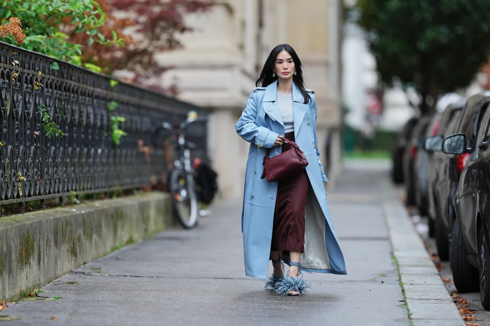 PARIS, FRANCE - NOVEMBER 07: Heart Evangelista wears long wavy black hair, a light blue leather double-breasted trench coat over a light gray knit top, burgundy wide-leg cropped trousers, blue feather-trimmed heeled sandals with ankle straps, a burgundy leather pouch bag, and turquoise drop earrings, during a street style fashion photo session, on November 07, 2025 in Paris, France. (Photo by Edward Berthelot/Getty Images)