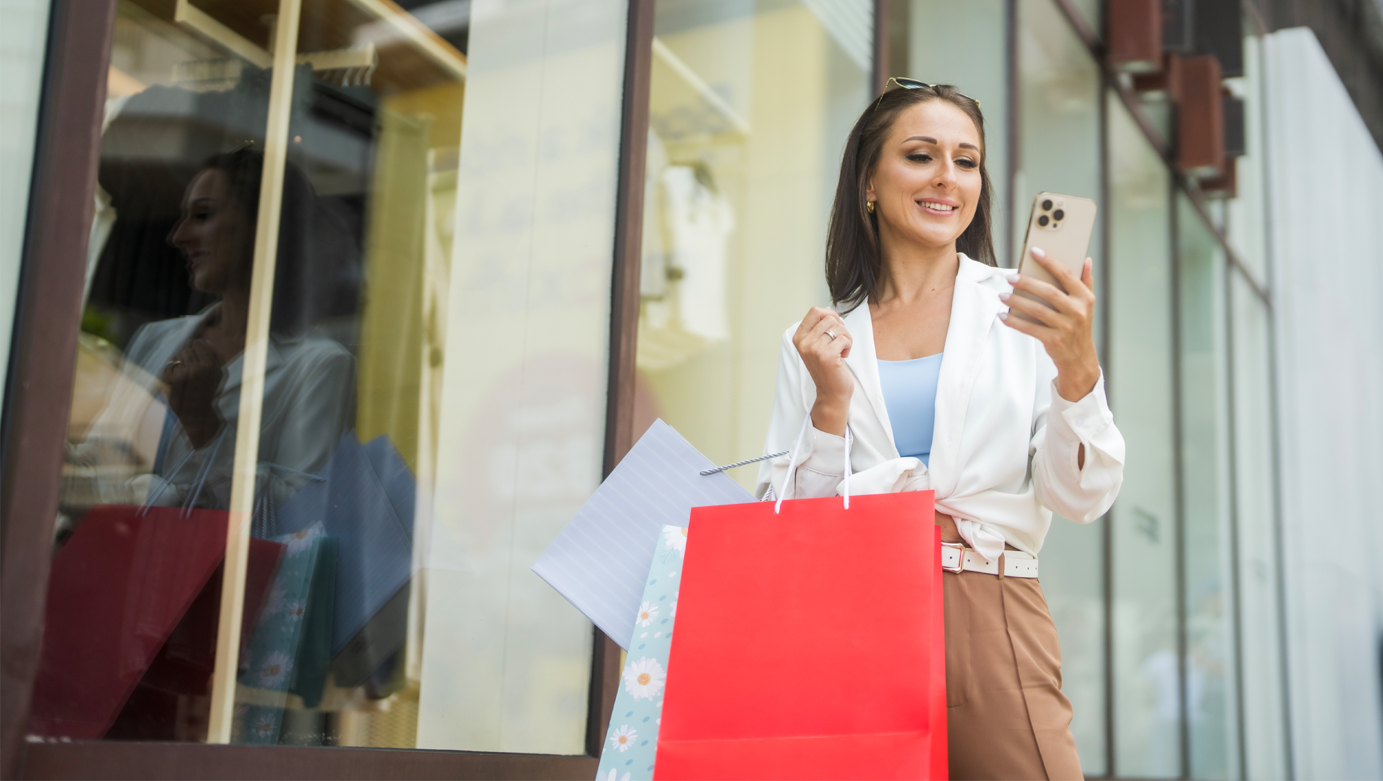 A joyful woman stands outside a store, holding shopping bags and looking at her smartphone, embodying a sense of satisfaction and modern lifestyle. Her expression suggests delight while shopping.