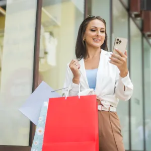 A joyful woman stands outside a store, holding shopping bags and looking at her smartphone, embodying a sense of satisfaction and modern lifestyle. Her expression suggests delight while shopping.