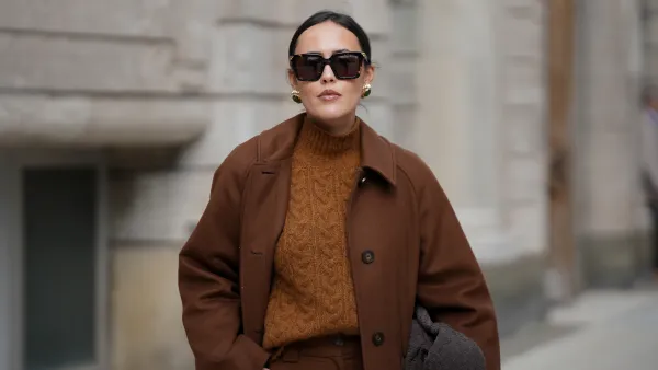 HAMBURG, GERMANY - OCTOBER 16: Laura Noltemeyer is seen wearing a long straight tailored wool coat in chocolate brown with button fastening by Sézane; high-waisted wide-leg pleated pants by Sézane; a textured cable knit sweater in a brown tone by Sézane; a suede asphalt grey shoulder bag with a woven leather structure by Les Visionnaires; square brown sunglasses with gold detailing by Bottega Veneta; gold statement earrings with a green stone detail by Mango; her dark hair is in a low ponytail on October 16, 2025 in Hamburg, Germany. (Photo by Moritz Scholz/Getty Images)