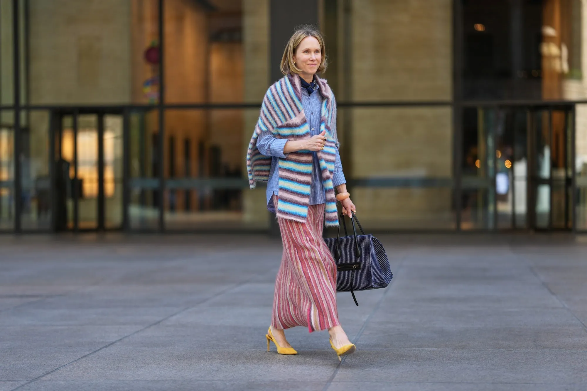 NEW YORK, NEW YORK - SEPTEMBER 15: A guest wears a shoulder-length blonde bob with a center part, a black pleated leather shoulder bag, a blue-and-white striped button-down shirt with rolled cuffs, a fuzzy horizontal-striped wrap in pastel blue, pink, white and brown draped over the shoulders, a long pleated skirt with vertical red, pink, cream and tan stripes, yellow suede pointed-toe slingback pumps shoes with a stiletto heel, outside Toteme, during New York Fashion Week, on September 15, 2025 in New York, New York (Photo by Edward Berthelot/Getty Images)