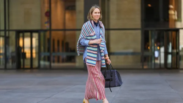 NEW YORK, NEW YORK - SEPTEMBER 15: A guest wears a shoulder-length blonde bob with a center part, a black pleated leather shoulder bag, a blue-and-white striped button-down shirt with rolled cuffs, a fuzzy horizontal-striped wrap in pastel blue, pink, white and brown draped over the shoulders, a long pleated skirt with vertical red, pink, cream and tan stripes, yellow suede pointed-toe slingback pumps shoes with a stiletto heel, outside Toteme, during New York Fashion Week, on September 15, 2025 in New York, New York (Photo by Edward Berthelot/Getty Images)