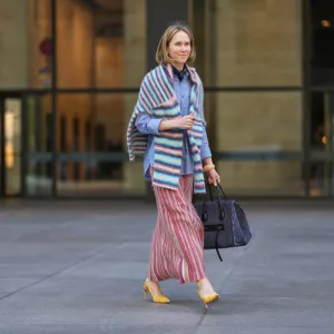 NEW YORK, NEW YORK - SEPTEMBER 15: A guest wears a shoulder-length blonde bob with a center part, a black pleated leather shoulder bag, a blue-and-white striped button-down shirt with rolled cuffs, a fuzzy horizontal-striped wrap in pastel blue, pink, white and brown draped over the shoulders, a long pleated skirt with vertical red, pink, cream and tan stripes, yellow suede pointed-toe slingback pumps shoes with a stiletto heel, outside Toteme, during New York Fashion Week, on September 15, 2025 in New York, New York (Photo by Edward Berthelot/Getty Images)