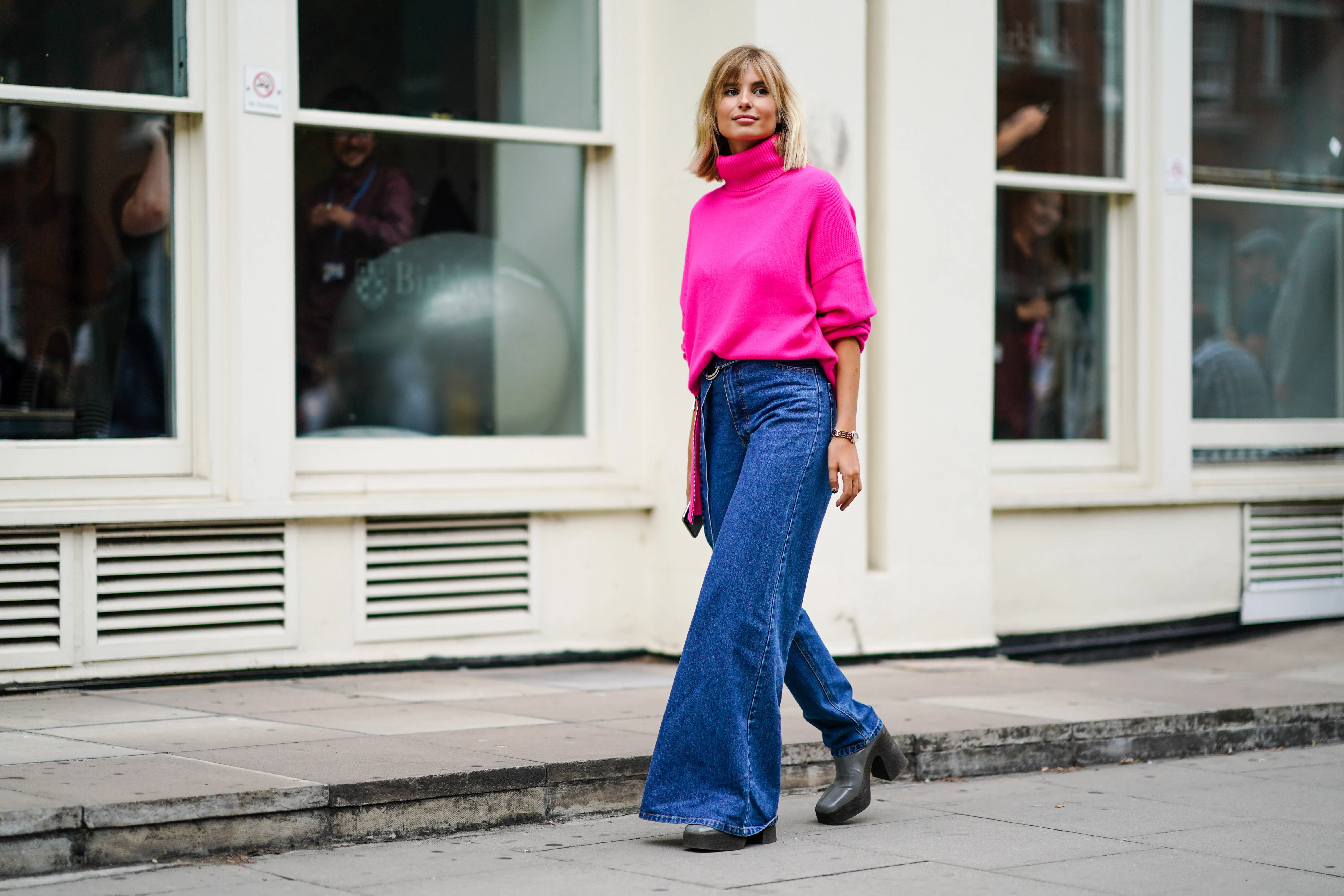 Xenia Adonts wears a neon pink turtleneck wool pullover, blue denim jeans pants with one flare leg, gray platform boots, during London Fashion Week September 2019 on September 16, 2019 in London, England.