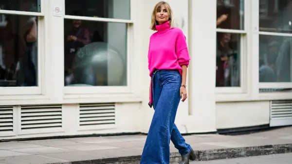Xenia Adonts wears a neon pink turtleneck wool pullover, blue denim jeans pants with one flare leg, gray platform boots, during London Fashion Week September 2019 on September 16, 2019 in London, England.