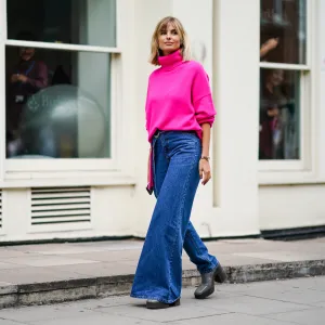 Xenia Adonts wears a neon pink turtleneck wool pullover, blue denim jeans pants with one flare leg, gray platform boots, during London Fashion Week September 2019 on September 16, 2019 in London, England.
