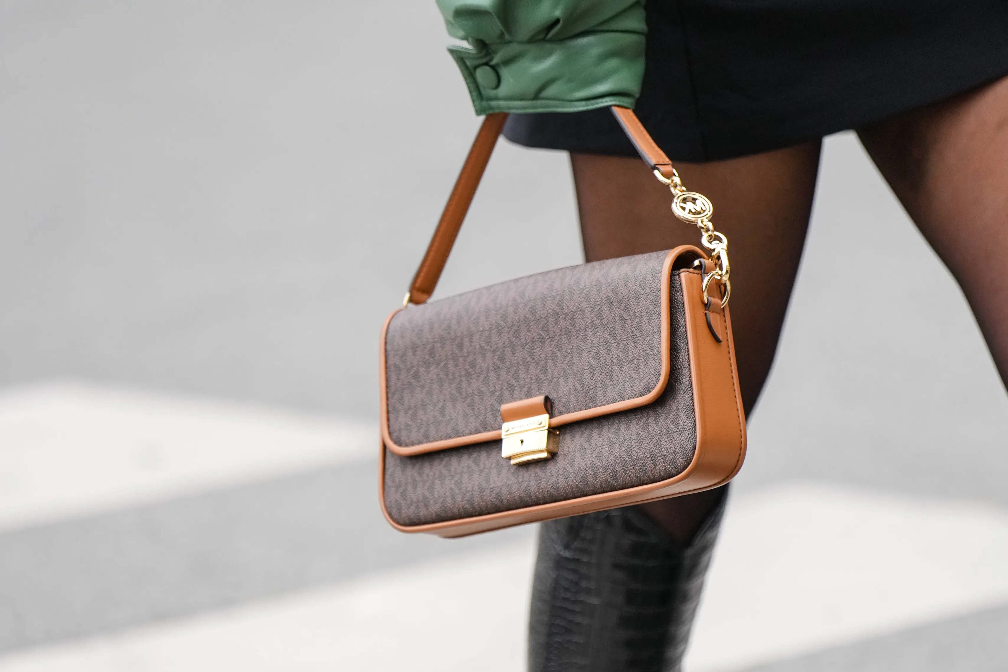 A guest wears a black turtleneck short tube dress, a khaki shiny leather oversized bomber coat, black tights, a brown monogram print pattern in coated canvas handbag from Mickael Kors, black shiny varnished leather crocodile print pattern pointed knees boots, outside Gauchere, during Paris Fashion Week - Womenswear F/W 2022-2023, on March 03, 2022 in Paris, France.