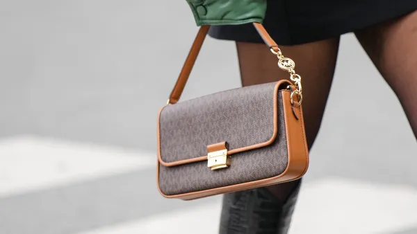 A guest wears a black turtleneck short tube dress, a khaki shiny leather oversized bomber coat, black tights, a brown monogram print pattern in coated canvas handbag from Mickael Kors, black shiny varnished leather crocodile print pattern pointed knees boots, outside Gauchere, during Paris Fashion Week - Womenswear F/W 2022-2023, on March 03, 2022 in Paris, France.