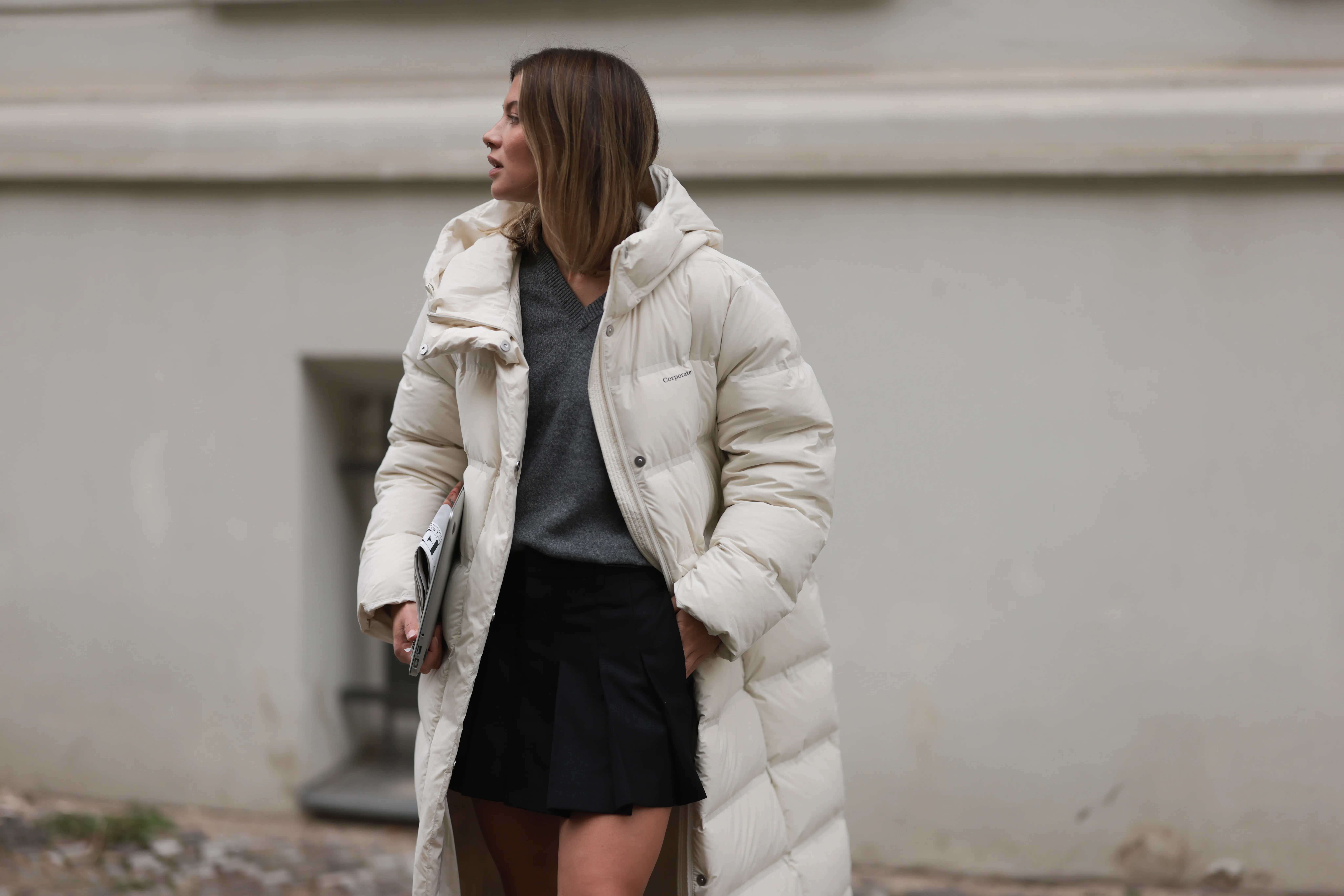Sophia Geiss is seen wearing a silver nose ring; an outfit from MO&Co. consisting of an ankle-length white quilted jacket, a grey pullover with a ribbed V-neckline underneath, and a black pleated mini skirt; while carrying a MacBook and a newspaper on September 26, 2024 in Berlin, Germany.