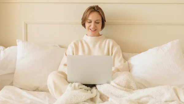 Smiling young woman in casual loungewear sitting on her bed working on her laptop computer