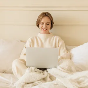 Smiling young woman in casual loungewear sitting on her bed working on her laptop computer